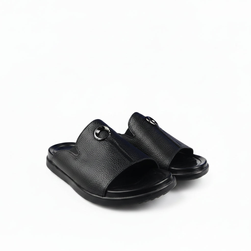 A pair of black pebbled leather slide sandals with a circular metal ring detail on the strap, shown on a clean white background.