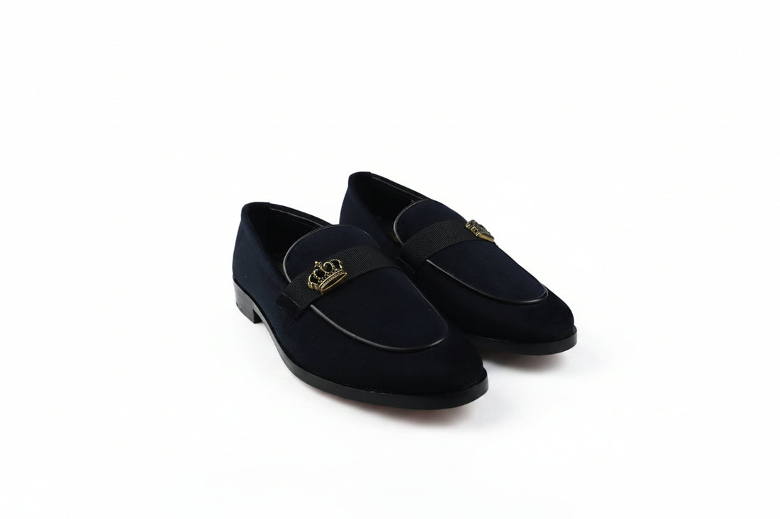 A pair of dark navy blue velvet loafers with a black strap across the vamp featuring a gold-tone crown emblem, set against a plain white background.