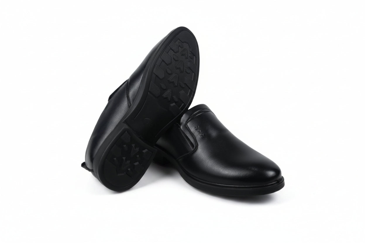 A pair of black  slip-on shoes, with one shoe standing upright to show the textured outsole and the other lying on its side to show the side profile, set against a plain white background.