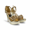 A pair of metallic gold high-heeled sandals with platform soles and ornate rhinestone-embellished straps, photographed against a plain light gray studio background.