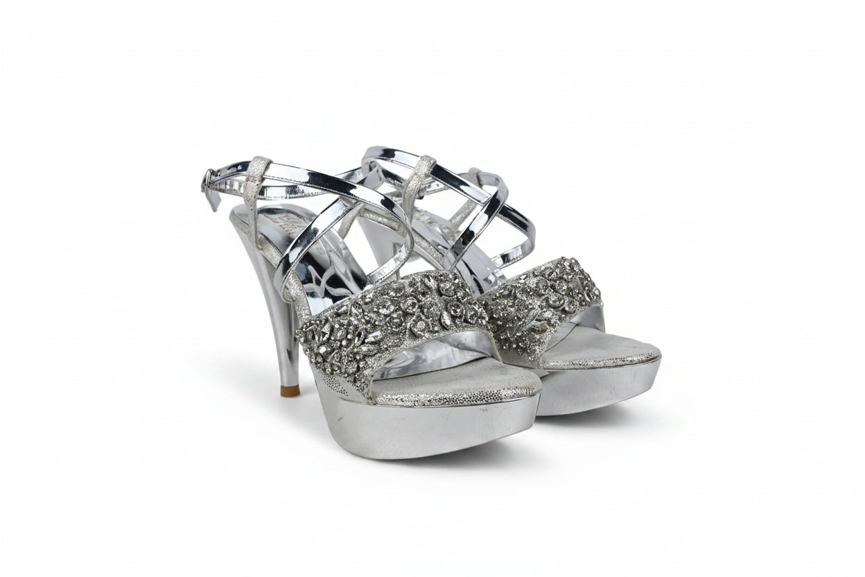 A pair of metallic silver high-heeled sandals with crystal-embellished straps and glittery platform soles, photographed on a light gray studio background.