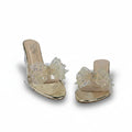 A pair of gold metallic slide sandals with clear straps adorned with large, embellished gold and silver rhinestone bows, set against a plain light gray background.
