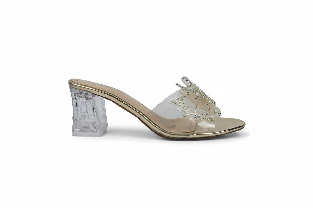 A single women's heeled mule sandal with a clear transparent block heel, a metallic gold footbed and trim, and a clear strap embellished with rhinestones, shown against a white studio background.