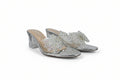 A pair of silver metallic mule sandals with clear transparent straps decorated with white and iridescent rhinestones, set against a light gray fabric background.