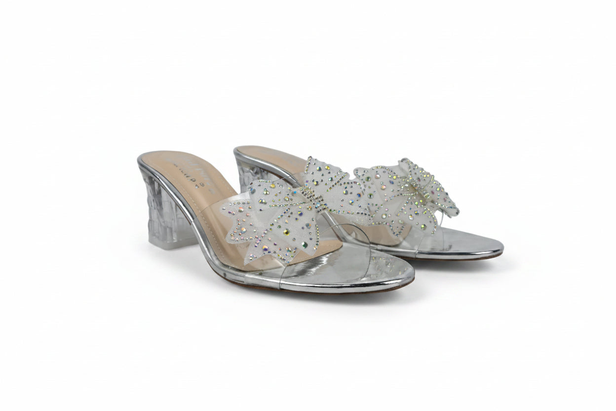 A pair of silver metallic mule sandals with clear transparent straps decorated with white and iridescent rhinestones, set against a light gray fabric background.