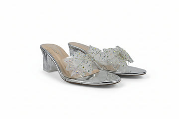 A pair of silver metallic mule sandals with clear transparent straps decorated with white and iridescent rhinestones, set against a light gray fabric background.