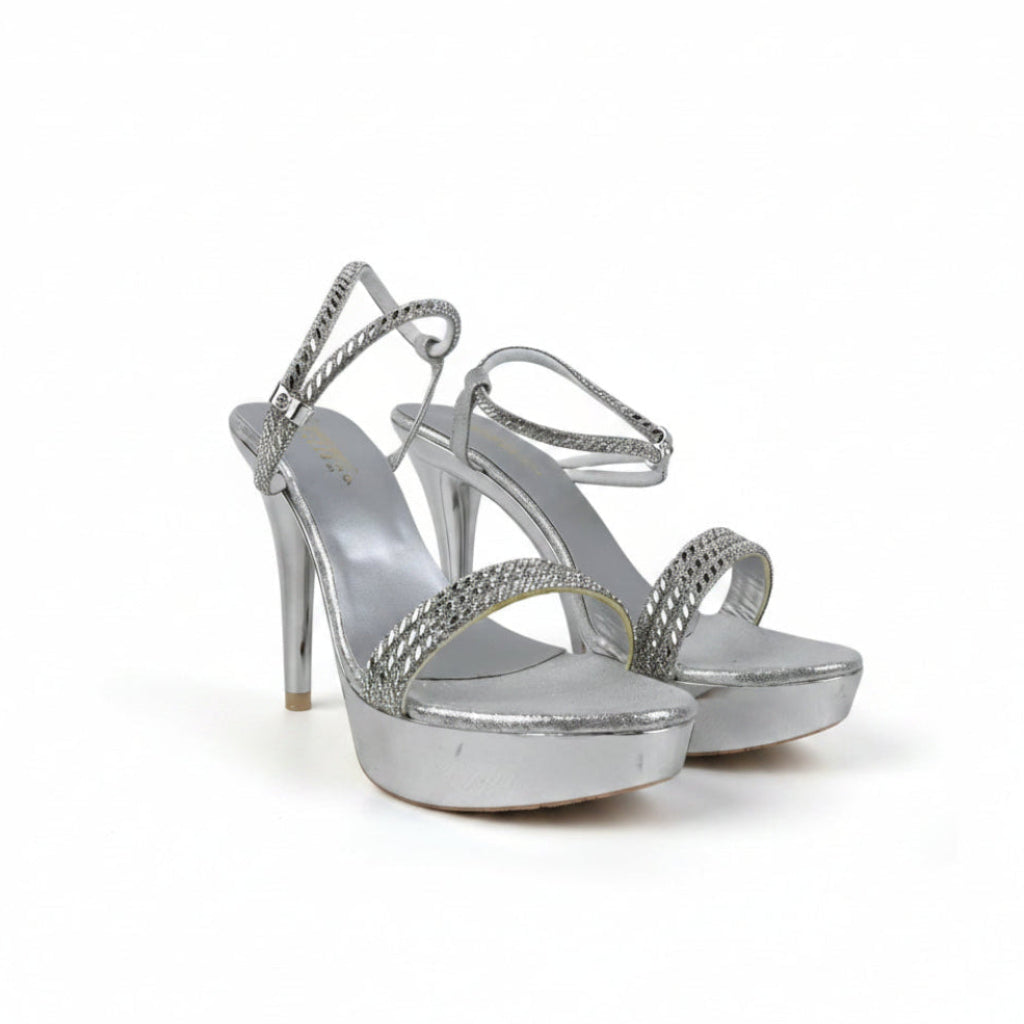 A pair of metallic silver high-heeled sandals with platform soles and braided strap details, photographed against a light gray textured background.