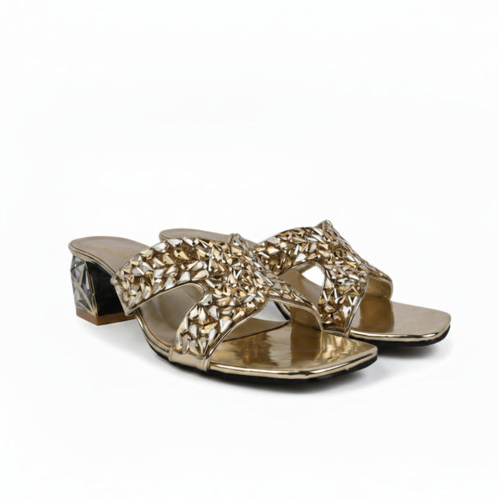 A pair of metallic gold heeled slide sandals with intricate rhinestone-embellished straps, displayed on a light gray textured surface.