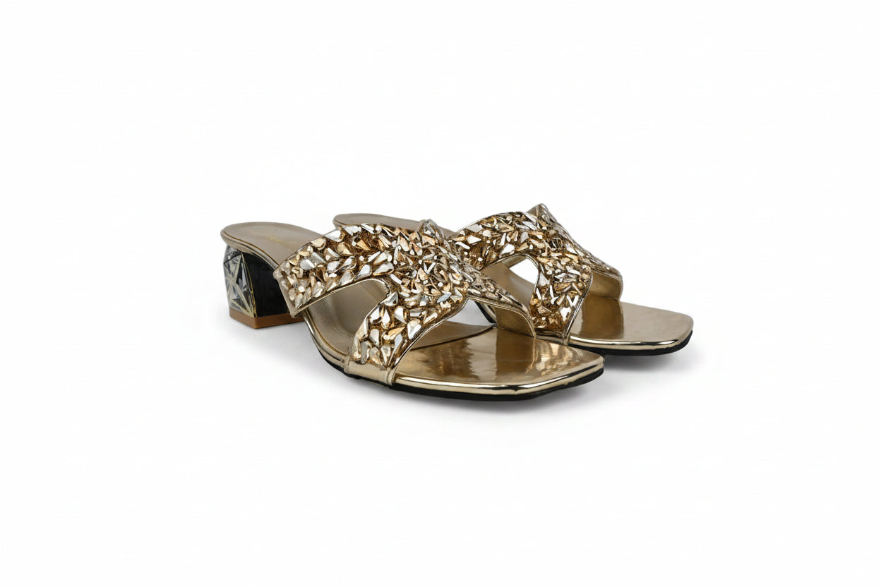 A pair of metallic gold heeled slide sandals with intricate rhinestone-embellished straps, displayed on a light gray textured surface.