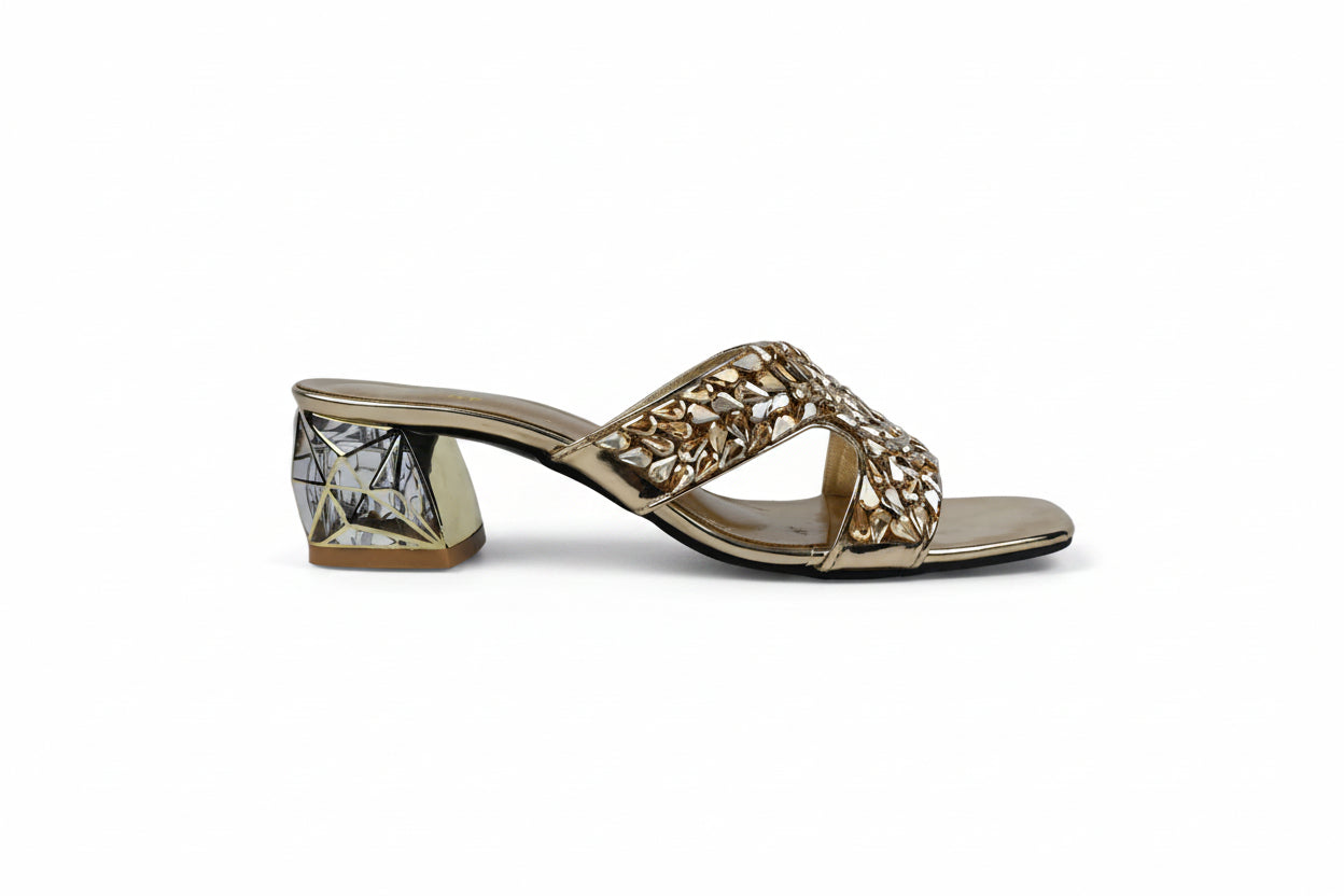 A single women's heeled mule sandal with a gold-tone metallic frame, a chunky block heel featuring a black and white geometric pattern, and a wide crisscross front strap embellished with gold rhinestones, shown against a light gray background.
