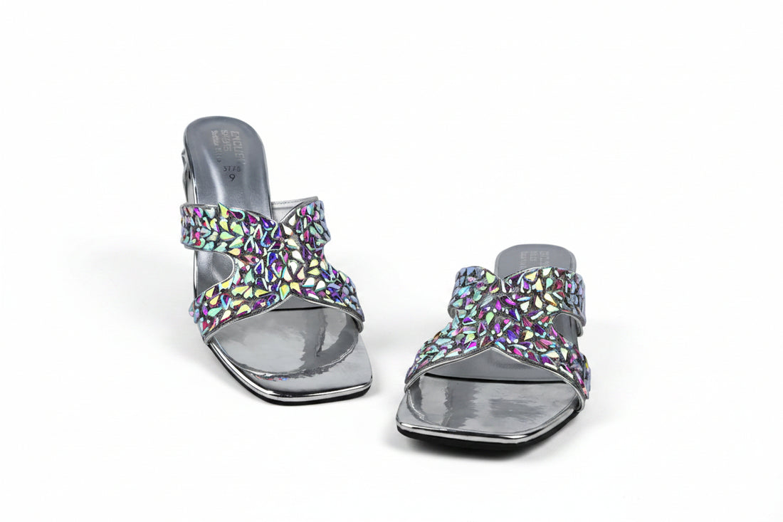 A pair of metallic gold heeled slide sandals with intricate rhinestone-embellished straps, displayed on a light gray textured surface.