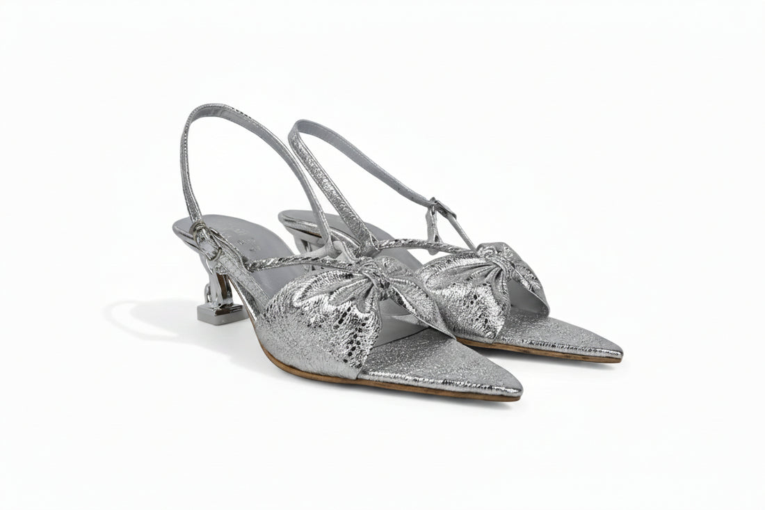 A pair of metallic silver slingback kitten heel sandals with pointed toes and large bow embellishments on the front, displayed against a light gray background.