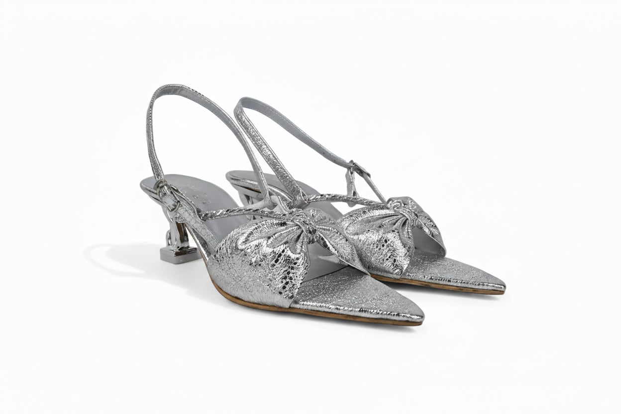 A pair of metallic silver slingback kitten heel sandals with pointed toes and large bow embellishments on the front, displayed against a light gray background.