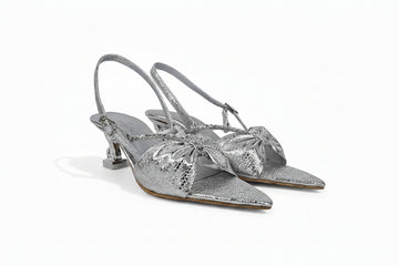 A pair of metallic silver slingback kitten heel sandals with pointed toes and large bow embellishments on the front, displayed against a light gray background.
