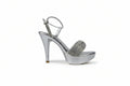 A single metallic silver high-heeled sandal with an ankle strap and a sparkling embellished toe strap, shown against a light gray fabric backdrop.