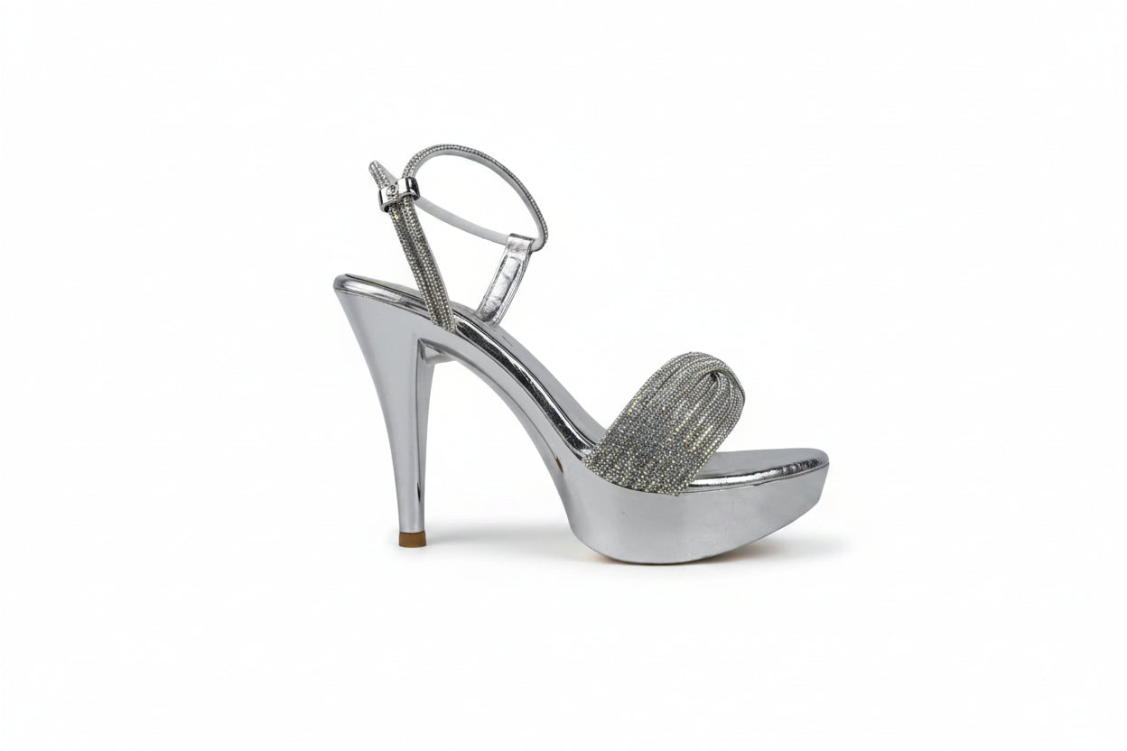 A single metallic silver high-heeled sandal with an ankle strap and a sparkling embellished toe strap, shown against a light gray fabric backdrop.