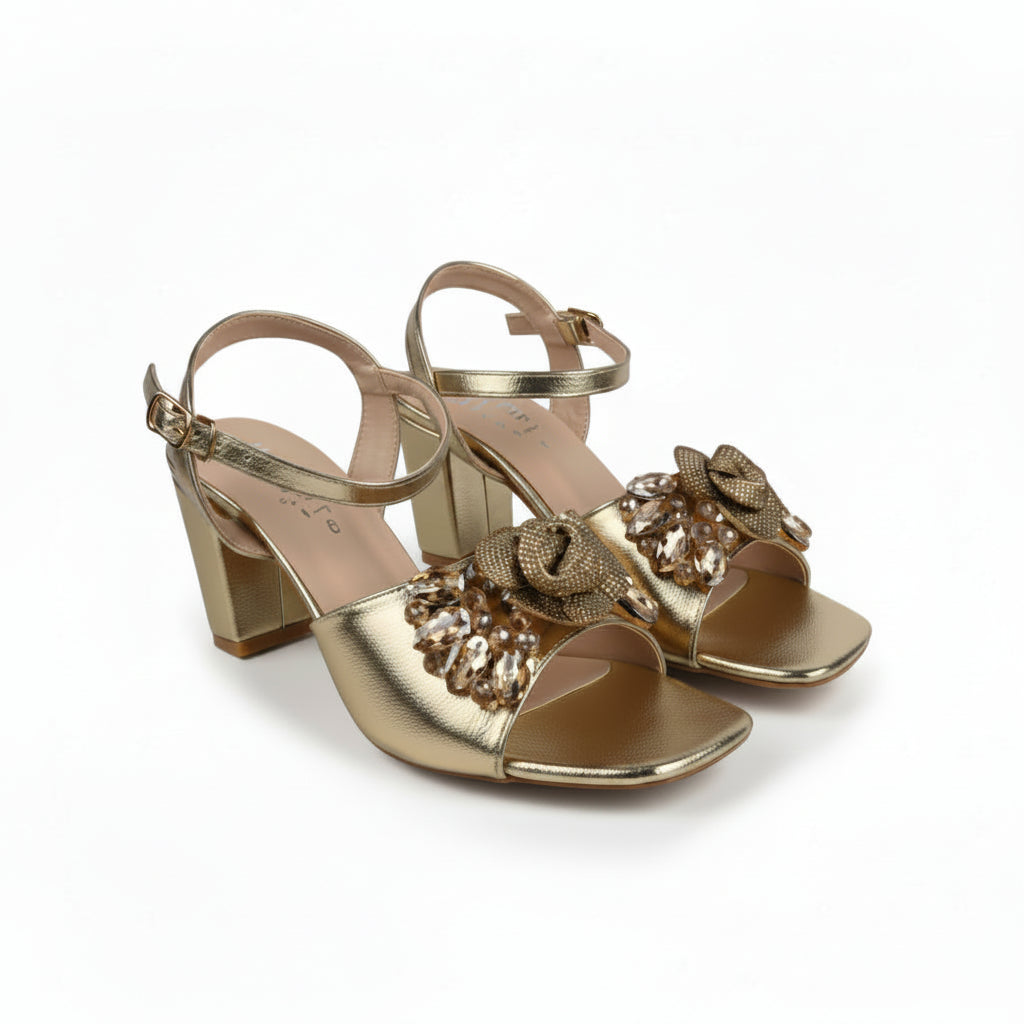 A pair of metallic gold heeled sandals with ankle straps and decorative bow embellishments on the front, displayed on a light gray studio background.