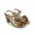 A pair of metallic gold heeled sandals with ankle straps and decorative bow embellishments on the front, displayed on a light gray studio background.