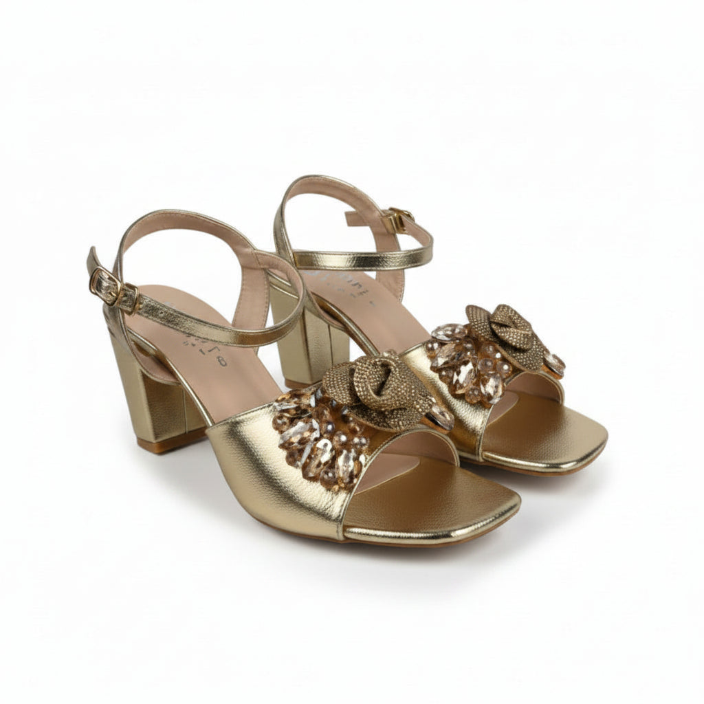 A pair of metallic gold heeled sandals with ankle straps and decorative bow embellishments on the front, displayed on a light gray studio background.