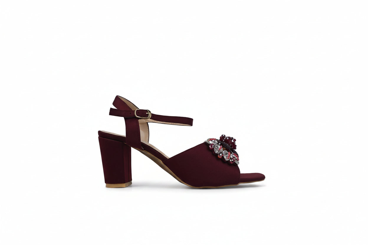 A single women's heeled sandal in deep burgundy with a block heel, ankle strap, and a jeweled floral embellishment on the toe strap, shown against a light gray studio backdrop.