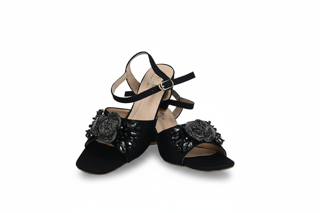 A pair of black open-toe slingback sandals with ankle straps and large black beaded floral embellishments on the toes, photographed against a white studio background.