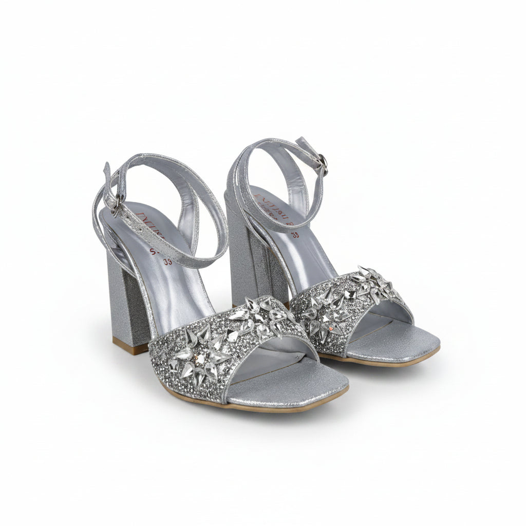 A pair of silver metallic high-heeled sandals with ankle straps and heavily embellished crystal-embellished toe straps, shown on a light gray studio background.