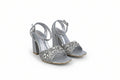 A pair of silver metallic high-heeled sandals with ankle straps and heavily embellished crystal-embellished toe straps, shown on a light gray studio background.