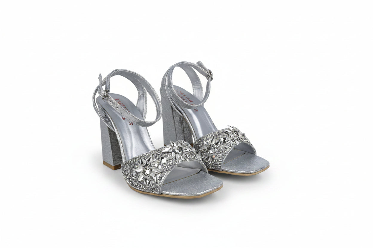 A pair of silver metallic high-heeled sandals with ankle straps and heavily embellished crystal-embellished toe straps, shown on a light gray studio background.