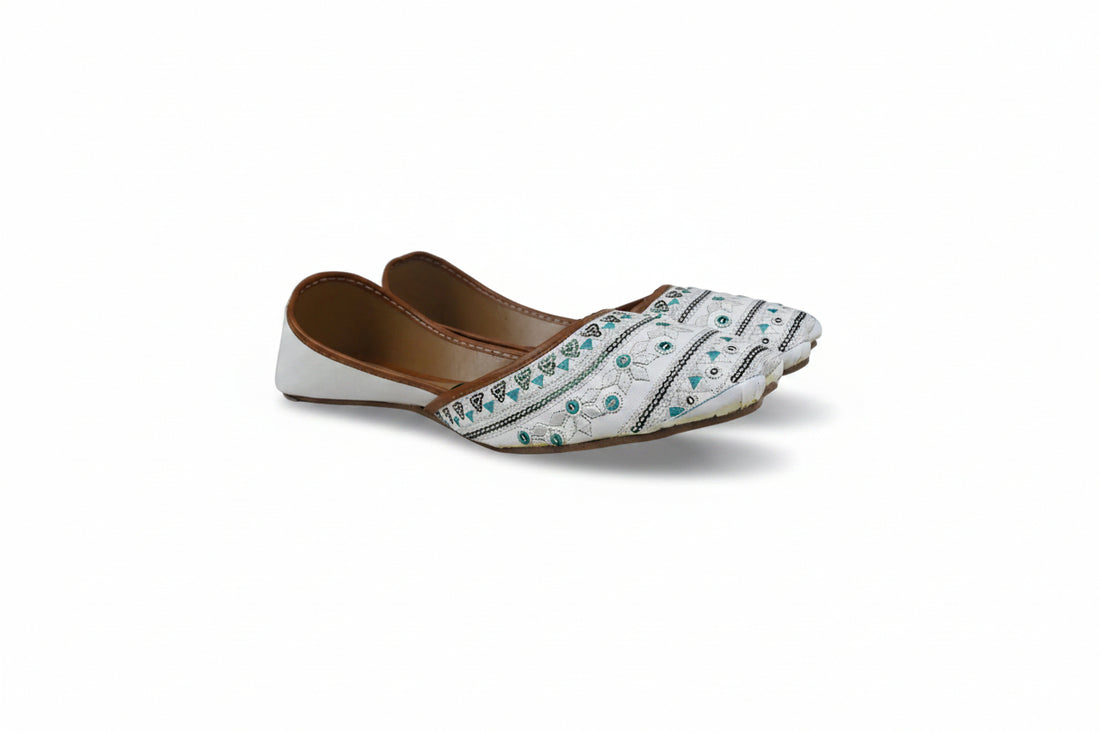 A pair of white flat jhumka-style shoes with brown leather trim and turquoise blue beadwork, displayed on a light gray fabric background.