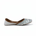 A single white flat shoe with brown leather trim and blue, black, and white geometric embroidery on the toe, shown against a light gray background.