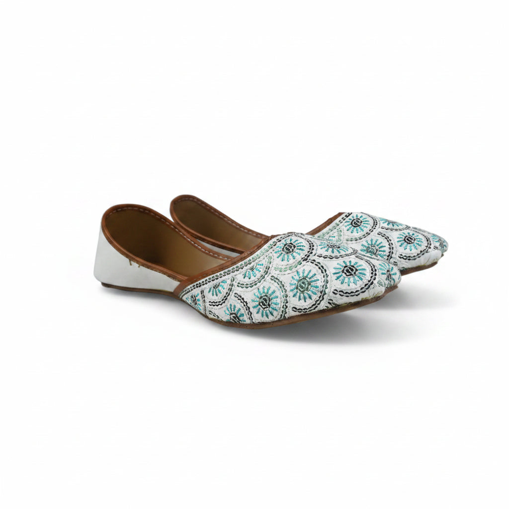 A pair of white pointed-toe flat shoes decorated with intricate blue, black, and white geometric embroidery, shown against a light gray background.