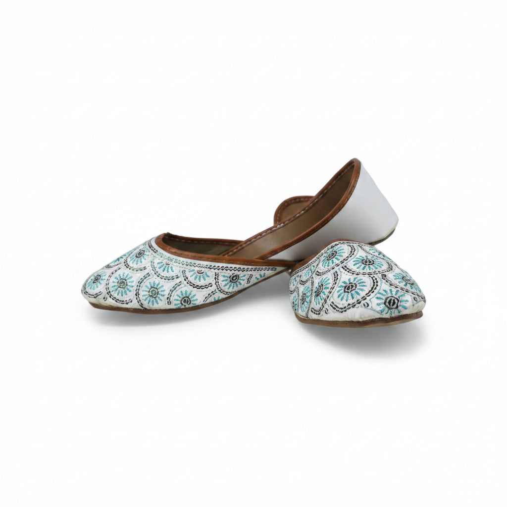 A pair of white pointed-toe flat shoes decorated with intricate blue, black, and white geometric embroidery, shown against a light gray background.