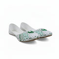 A pair of white flat shoes with intricate green and turquoise beadwork and sequins on the toe caps, photographed on a light gray surface.