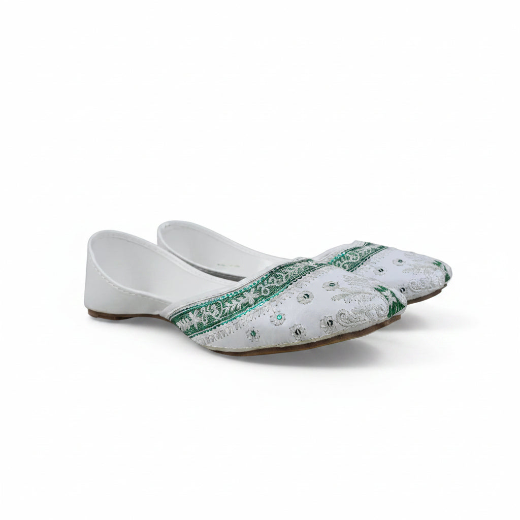 A pair of white traditional flat shoes with green embroidered borders and silver mirror work, photographed on a light gray fabric backdrop.