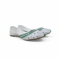 A pair of white traditional flat shoes with green embroidered borders and silver mirror work, photographed on a light gray fabric backdrop.