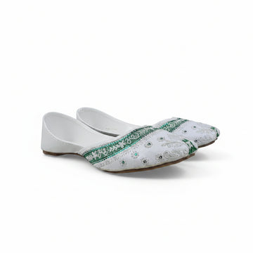 A pair of white traditional flat shoes with green embroidered borders and silver mirror work, photographed on a light gray fabric backdrop.