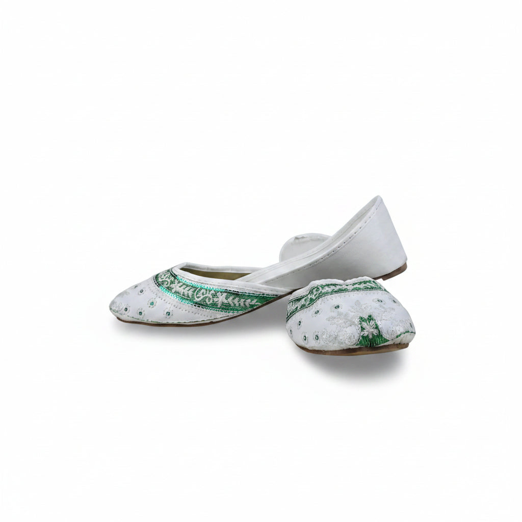A pair of white traditional flat shoes with green embroidered borders and silver mirror work, photographed on a light gray fabric backdrop.