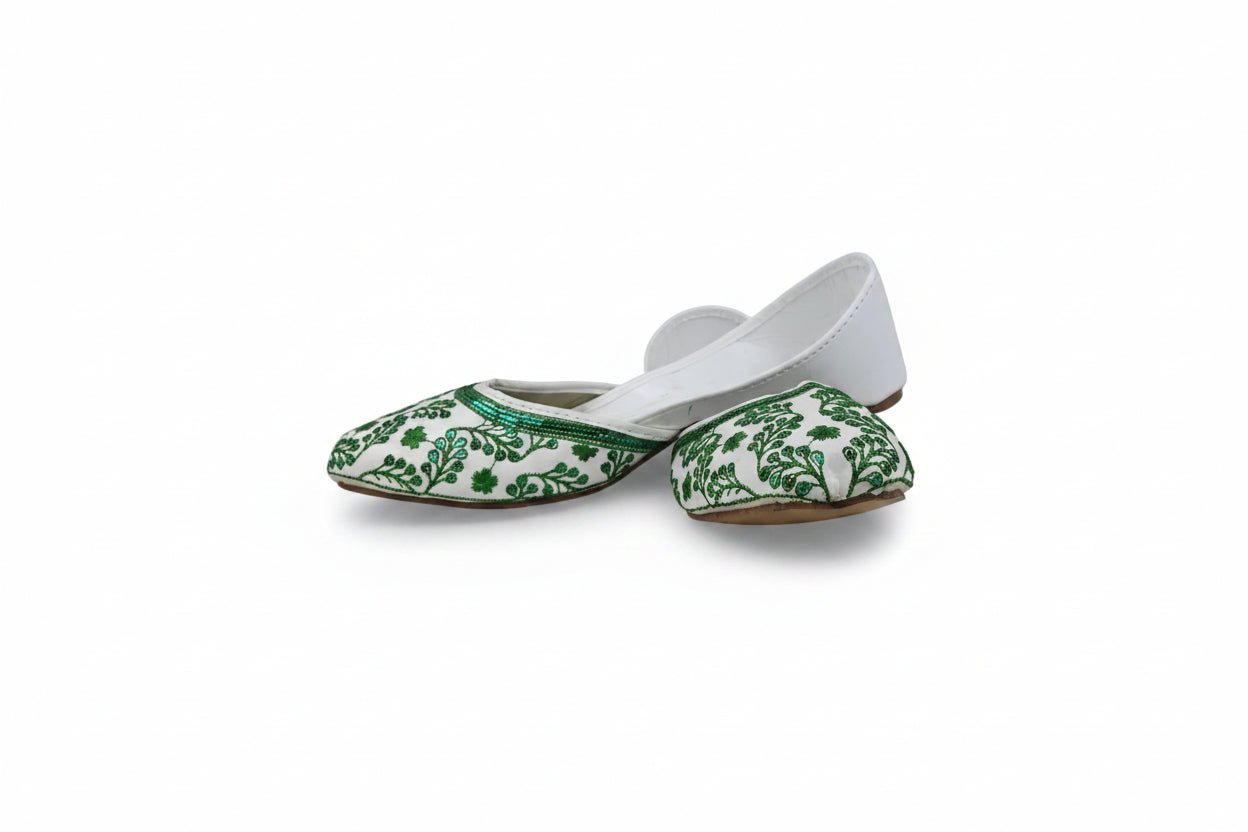 A pair of white flat shoes with green floral embroidery on the pointed toes, photographed against a light gray fabric backdrop.