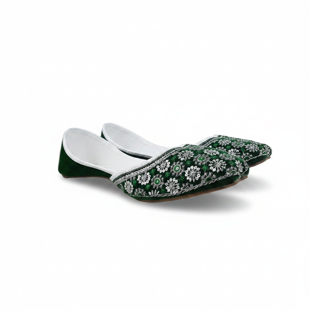 A pair of green velvet traditional flat shoes with intricate white floral embroidery and silver mirror work, displayed on a light gray fabric background.
