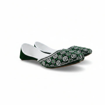 A pair of green velvet traditional flat shoes with intricate white floral embroidery and silver mirror work, displayed on a light gray fabric background.