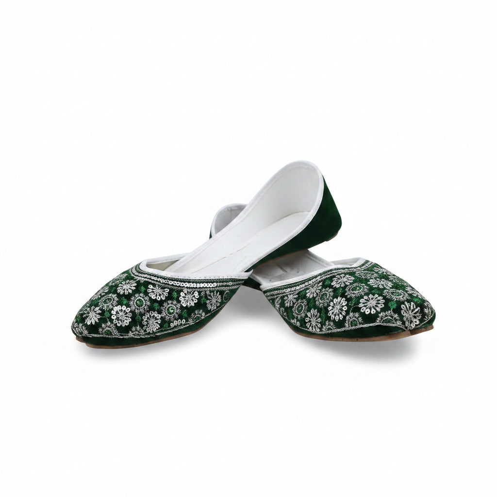 A pair of green velvet traditional flat shoes with intricate white floral embroidery and silver mirror work, displayed on a light gray fabric background.