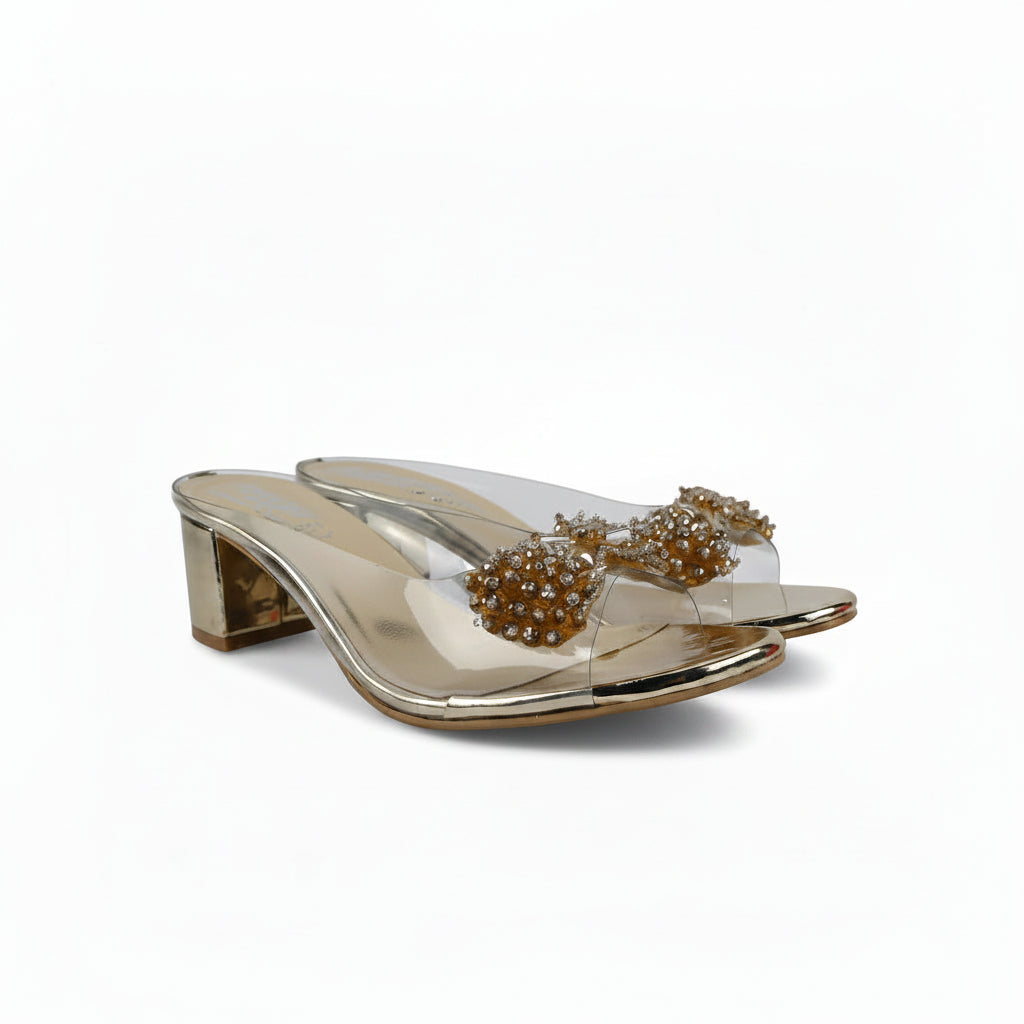 A pair of metallic gold heeled mule sandals with clear straps and large, beaded gold bows on the toes, set against a plain white background.