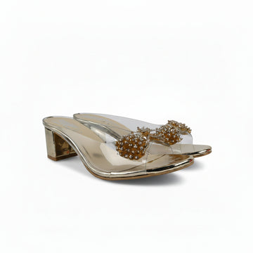 A pair of metallic gold heeled mule sandals with clear straps and large, beaded gold bows on the toes, set against a plain white background.