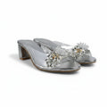 A pair of metallic silver heeled mule sandals decorated with large crystal flower embellishments on the front, set against a light gray fabric backdrop.