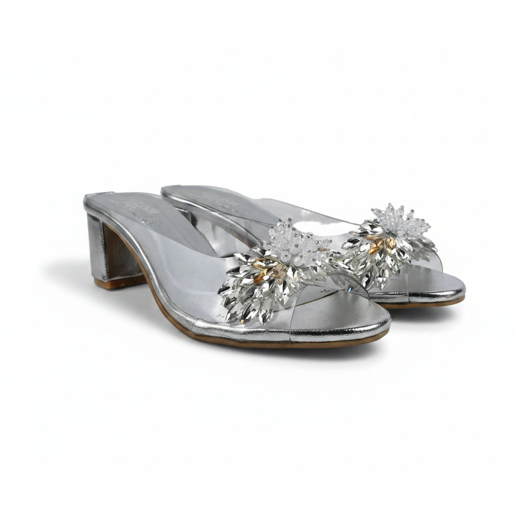 A pair of metallic silver heeled mule sandals decorated with large crystal flower embellishments on the front, set against a light gray fabric backdrop.