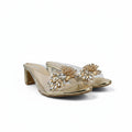A pair of gold heeled mule sandals with clear straps adorned with large gold and clear rhinestone floral embellishments, set against a white fabric backdrop.