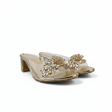 A pair of gold heeled mule sandals with clear straps adorned with large gold and clear rhinestone floral embellishments, set against a white fabric backdrop.