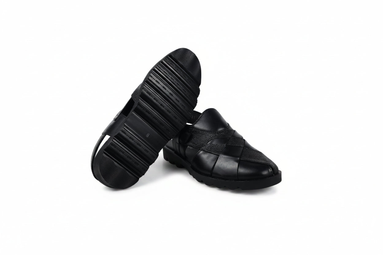A pair of black leather men's loafers, with one shoe standing upright to show the textured sole and the other lying flat to display the woven strap pattern, set against a white studio backdrop.