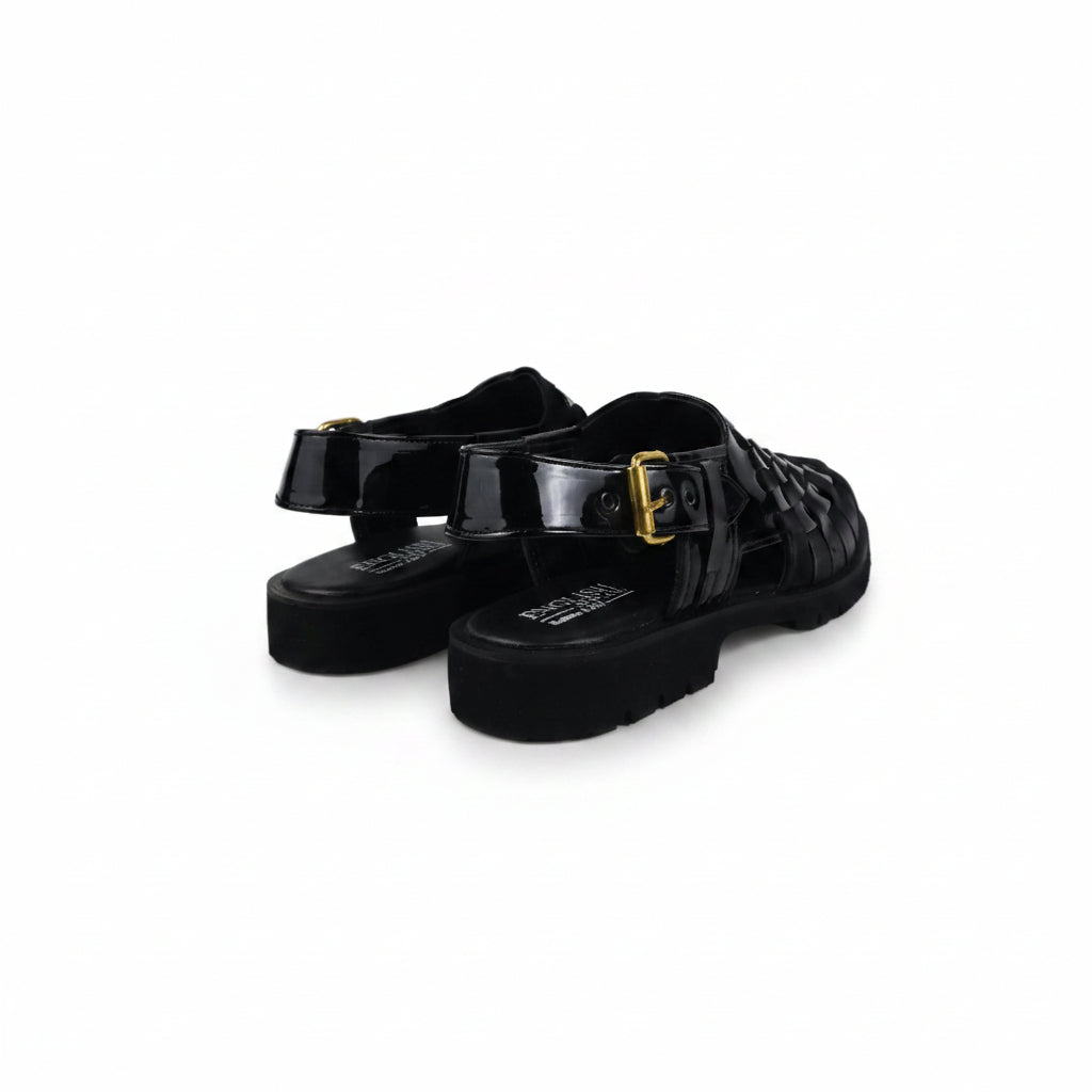 A pair of black patent leather mule sandals with buckle straps and chunky black soles, photographed on a white studio background.
