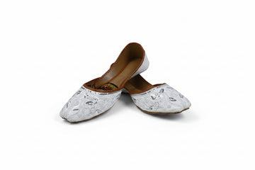 A pair of white embroidered flat shoes with brown insoles, photographed on a light gray studio background.